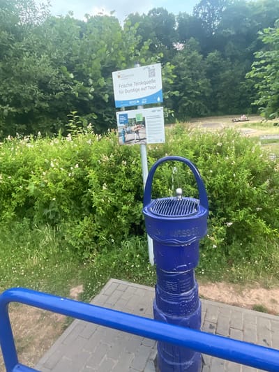 In Neukirchen, Germany, a vibrant blue public drinking fountain stands prominently on a paved area, its top section adorned with the inscription "Trink Wasser." Water gently streams from a silver spout into a basin. The fountain is surrounded by lush green foliage, including flowering bushes and trees, creating a natural and refreshing ambiance. A blue metal railing is visible in the foreground, and a signpost is positioned behind the fountain, displaying information in German, including "Frische Trinkquelle für Durstige auf Tour." In the background, people can be seen in the distance, suggesting a recreational area or park. The overall scene captures a moment of public amenity and natural beauty in the city.