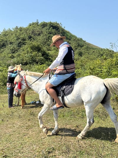 The image shows a man riding a white horse in a field in Bắc Hà, Vietnam. The man is wearing a straw hat, a white shirt, a black vest with colorful patterns, and light blue shorts. He is holding the reins of the horse. Another man is standing beside a horse with traditional decoration. The background features green vegetation and a clear blue sky. The scene conveys a rural setting and possibly a cultural activity or festival involving horses and traditional attire.