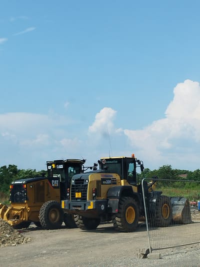 Two construction vehicles are parked on a gravel surface under a bright blue sky with scattered clouds. On the left is a yellow Caterpillar grader with the "CAT" logo visible. To its right is a yellow Komatsu wheel loader, identified by the "KOMATSU" branding and "WA320" model number. Both vehicles are large and appear to be stationary. In the background, there is a line of green trees and some structures. A metal fence with some bricks at its base is in the foreground, partially obscuring the wheel loader. The scene suggests a construction site in Bragadiru, Romania.