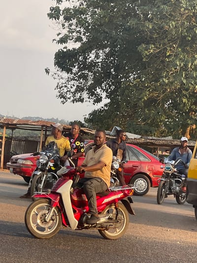 Several people on motorcycles are in the street. One person in a tan shirt on a red motorcycle looks at the camera. Another person is behind them with a colorful shirt. A few cars and a tree are in the background.