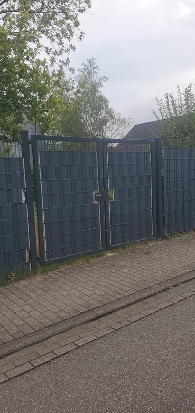 The image shows a gray metal gate with dark gray screening. The gate is divided into two panels and has a latch mechanism. The gate is set against a backdrop of green trees and a cloudy sky. In the foreground, there is a paved walkway made of interlocking concrete blocks, adjacent to a darker asphalt road. The scene appears to be in an outdoor urban or suburban environment. There is no visible indication of the specific location within Lauf, Germany, nor are there any people present in the image.