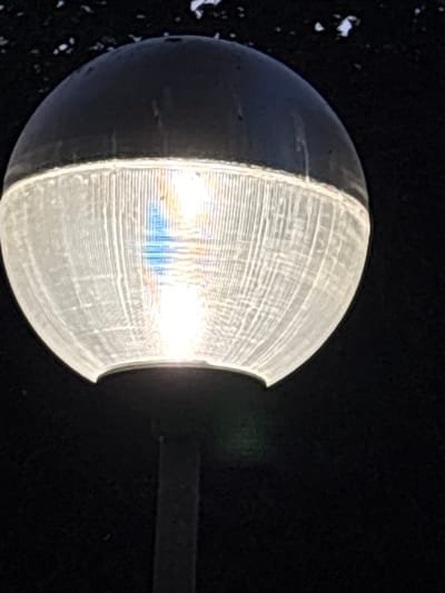 A close-up, low-angle shot shows a black spherical light fixture with a clear, ribbed plastic globe housing a bright light bulb. The fixture is mounted on a dark pole, and the background is dark, suggesting it is nighttime. The image appears to have been taken outdoors in the city of Coslada, Spain, as indicated by the context provided. There are no people or specific actions depicted in the scene.