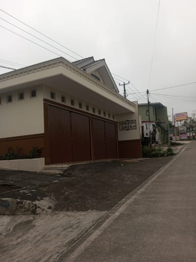 A building with a brown and white facade has closed brown metal doors. A sign reads "PONDOK DAFKO". A grey concrete sidewalk is in front. Across the street, there is another building and a red and white "Alfamart" sign. Utility lines crisscross the cloudy sky above. The weather looks overcast.