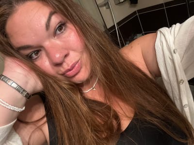 A woman poses for a selfie indoors. She is fair-skinned, with brown eyes and long brown hair. She is wearing a black tank top and a white cardigan or shirt. On her left wrist, she has a metal bracelet and a beaded bracelet. She is also wearing a beaded necklace with a small charm. The background includes black and white tiles, potentially in a bathroom setting. Based on the prompt, the location is Bijela, Montenegro, but there are no visible elements within the image that confirm this.