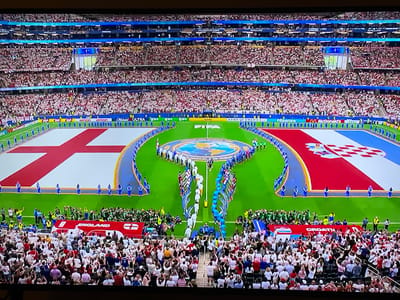 The image depicts a large, outdoor stadium filled with a vast crowd of spectators. The field of play is marked with large flags representing England and Croatia, with a circular emblem at the center of the field. Numerous individuals, likely participants, are lined up on the field, some holding flags. The spectators are densely packed into tiered seating that surrounds the field, with a significant number of them holding up flags and appearing to be cheering or engaged in a celebratory event. The overall atmosphere suggests a sporting event or a major public gathering. The location is identified as Florești, Romania.