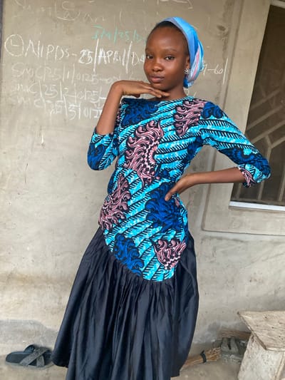 A young woman poses in a patterned blue, pink, and black top paired with a long black skirt and matching sandals. She wears a light blue headscarf and earrings. The backdrop is a wall scribbled with chalk, featuring a window above. A broom rests in the corner. The photograph is taken from a slightly elevated angle and turned sideways.