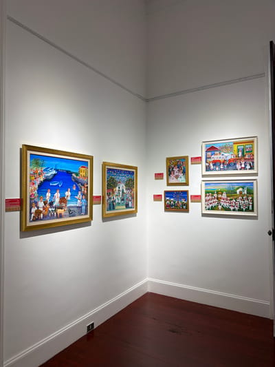 This is an indoor shot of a gallery displaying several colorful, folk art paintings on a white wall. The paintings are arranged on two walls, meeting at a corner, with a dark wooden floor visible in the foreground. 

On the left wall, a large, framed painting depicts a harbor scene with people, boats, and farm animals. To the right of this, a smaller, framed painting shows a group of people in a courtyard, possibly at a festival, with colorful banners overhead. Further to the right, on the adjacent wall, a series of smaller paintings are displayed. One shows children in formal attire, another a windmill with figures, and two larger paintings show communal gatherings. One of these larger paintings features a scene with a white truck and children, while the other depicts a wedding or religious procession in front of a church. Each painting has a small red informational plaque beneath it. The overall impression is one of a vibrant exhibition of traditional art. 

The information provided about the paintings and their location suggests this scene is from the museum of history and anthropology, Casa de los Balcones, in La Orotava, Spain.