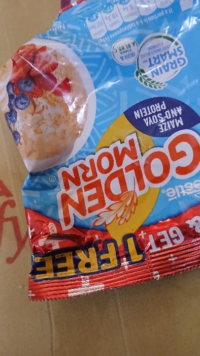 The image displays a slightly angled, close-up view of a Nestle Golden Morn food product package, positioned in Isasi, Nigeria. The package is a flexible, blue and red plastic pouch. The upper section is predominantly blue, featuring the "GOLDEN MORN" brand name in large orange letters, along with text identifying it as "MAIZE AND SOYA PROTEIN" and highlighting its "GRAIN SMART" qualities, including "IRON & VITAMIN A, B1, B5, C". An image of a bowl of cereal topped with sliced strawberries and blueberries is visible on the upper left. Nutritional information in white text is partially legible at the top. The lower section of the package is red and crinkled, with the phrase "GET 1 FREE" printed in yellow and white. The entire package rests on a light brown, textured surface, possibly cardboard, with faint red, indistinct markings visible on the left side of the background. No individuals are present in the scene.