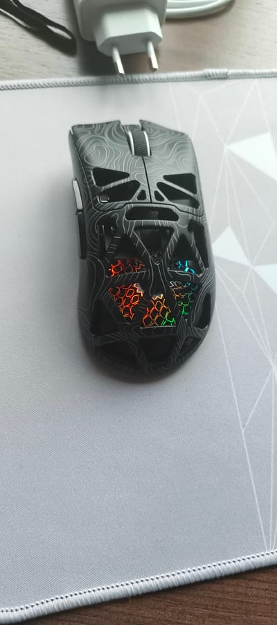 The image shows a close-up, top-down view of a black gaming mouse with a white topographic-style pattern. The mouse is positioned on a light gray mousepad with a geometric pattern. In the upper portion of the image, out of focus, is a white electrical adapter with two prongs and a white cable. The mouse has RGB lighting visible through cutouts, displaying a gradient of red, orange, yellow, green, and blue light. The scene is set indoors, on a wooden surface that is partially visible. There are no people present in the image. The location of Aktanyshbash, Russia is not depicted or inferable from the image.