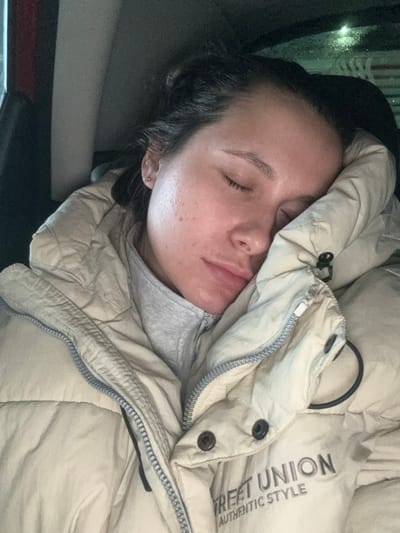 The image depicts a female individual sleeping, located in Vorsha, Russia. She is situated within an enclosed, dimly lit space, consistent with a vehicle interior, evident by dark surfaces and subtle reflections in the upper right background. The individual has closed eyes, dark hair pulled back, and a fair complexion with minor freckles visible on her left cheek. A small silver stud earring is present in her left earlobe. She is wearing a light-beige puffer jacket over a grey zippered garment. The jacket features the text "STREET UNION AUTHENTIC STYLE" on the right front section. Her head is angled to the right, resting against the jacket.