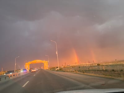 The image depicts a scenic drive under a dramatic sky, likely during a transition between day and evening. The scene is set on a highway in Satbayev, Kazakhstan, with a prominent yellow archway spanning the road bearing the text "SAT SAPARI" in red letters. This archway serves as an entrance or landmark.
The sky is filled with dark, ominous clouds, suggesting recent or impending rain. However, a beautiful double rainbow has emerged, arcing across the sky and casting a warm, colorful glow on the horizon. This juxtaposition of stormy clouds and a vibrant rainbow creates a visually striking and somewhat emotional atmosphere, perhaps evoking feelings of hope or awe.
Several vehicles are visible on the highway, including cars and what appears to be a truck or bus, indicating regular traffic. Streetlights are also present, some of which are illuminated, suggesting that the time of day is either early morning or late afternoon/early evening, as the natural light fades. In the background, there are power lines and what might be industrial or infrastructure structures, characteristic of a developed area. The landscape is predominantly flat and arid, with dry grass and sparse vegetation lining the sides of the road. The overall impression is of a journey through a vast landscape, marked by both the power of nature and human infrastructure.