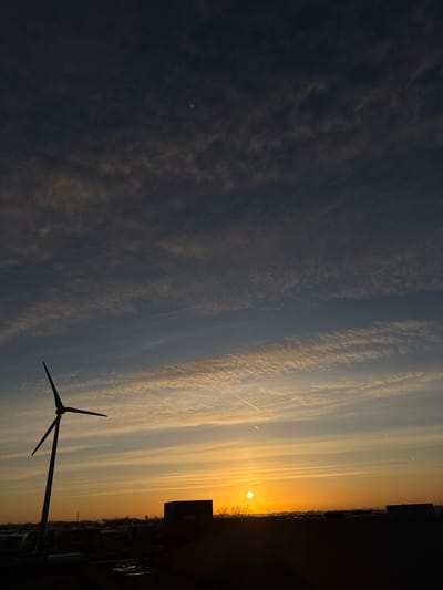 The image displays a twilight scene over an urban-industrial landscape in Naaldwijk, Netherlands, with the sun positioned low on the horizon. The upper two-thirds of the frame are dominated by the sky, exhibiting a vertical color gradient from deep blue-grey at the top, transitioning through lighter blue bands, to an intense orange and yellow glow at the horizon. The sun is visible as a bright, distinct orange-yellow circular disc situated low and slightly left of the image's horizontal center. Various cloud formations, including scattered altocumulus and cirrocumulus, are present across the sky. Several linear contrails traverse the mid-sky, with one distinct contrail extending diagonally from the upper-left to the mid-right. A faint, circular white celestial body, consistent with the moon, is discernable in the upper-left quadrant.
The lower third of the image comprises dark silhouettes of man-made structures. A large, three-bladed wind turbine stands prominently on the left, its dark mast extending from the lower-left corner. To the right of the wind turbine, a series of dark, rectangular industrial buildings and rooftops form the horizon line, notably including a large, blocky structure featuring a dark, recessed section. At ground level, dark, reflective patches of material, possibly wet ground or standing water, are visible. The faint outline of a white box-trailer truck can be identified near the base of the wind turbine. No human figures or explicit human activities are depicted within the scene.