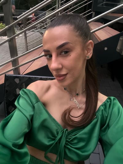 A woman is seated in a black chair in an outdoor setting. She is wearing a green off-the-shoulder top with puffy sleeves and a tie detail in the front. She has a silver necklace with a nameplate and clover pendants. She is also wearing small green clover stud earrings. The background shows a staircase with metal railings and wooden steps, and people seated at tables in the distance. The image appears to be taken in the city of Petrich, Bulgaria, likely at an outdoor restaurant or cafe.