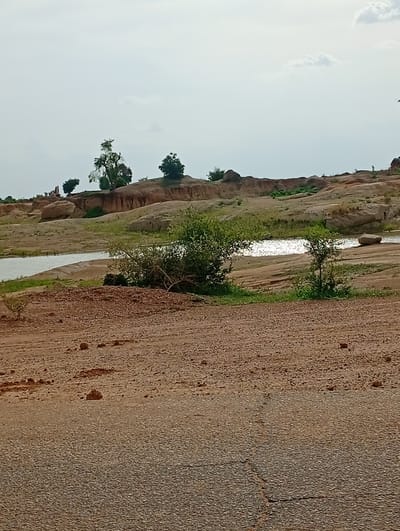 This is a full, eye-level, medium shot of a dusty, unpaved road in Shuwa, Nigeria. The road occupies the foreground and a portion of the midground of the image. Beyond the road, there is a small body of water, reflecting the bright, overcast sky. The water is bordered by a rocky, dry landscape with sparse vegetation, including several trees and bushes. In the background, a cluster of people can be seen standing on a rocky outcrop near a few trees. The sky is mostly clear with a few scattered clouds.