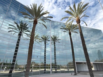 This is a photograph of the exterior of a modern office building in Garden Grove, California, United States of America. The building features a facade of large, reflective glass panels that mirror the sky and surrounding environment. Several tall palm trees are visible in the foreground, with their fronds extending towards the sky. The reflections in the glass show palm trees, clouds, and a distant cityscape. The sky is partly cloudy with a mix of blue sky and white clouds. The ground in front of the building appears to be a paved plaza area. A few pedestrians are visible in the reflection on the ground level, walking along a pathway. The overall impression is one of a contemporary urban environment with a touch of tropical landscaping.