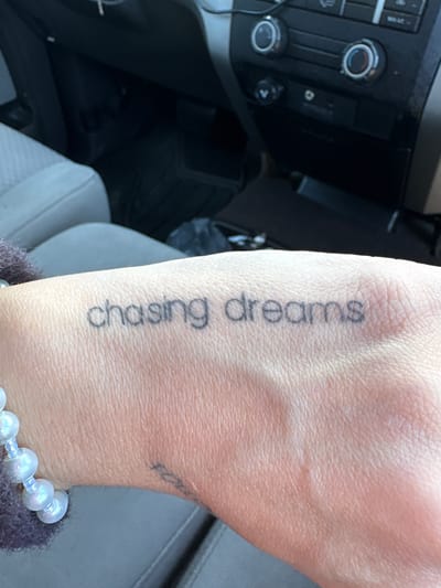 A person's hand is visible, featuring a tattoo that reads "chasing dreams" in a sans-serif font oriented vertically along the wrist and forearm. The hand also has a bracelet of iridescent white beads. The setting appears to be the interior of a vehicle, with a dashboard visible in the background. The dashboard includes climate control knobs and buttons. The seats are gray and appear to be upholstered in a cloth or synthetic material. The location is likely within Long Beach, California, USA as requested, but there are no visual cues to definitively identify a specific landmark or street within Long Beach.