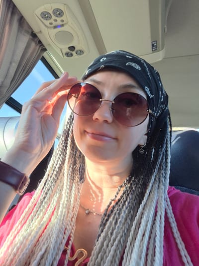 A woman with gray and white braided hair is wearing a black bandana and sunglasses, with her hand touching her sunglasses. She is wearing a pink shirt, a necklace, and an earring. The image appears to be taken inside a vehicle, possibly a bus, with a view of the sky and clouds through a window. The location in the city of Verkhniye Tatyshly, Russia is not discernible from the image itself.