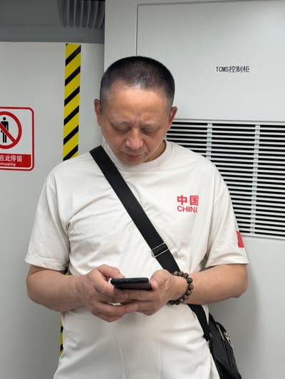 A middle-aged East Asian man with short, dark hair is standing indoors, looking down at a smartphone he is holding in his hands. He is wearing a white t-shirt with red Chinese characters and the word "CHINA" printed on it. A black strap for a bag is slung across his chest. He is also wearing a bracelet made of wooden beads on his left wrist. The man is standing in front of a white wall with black and yellow diagonal stripes on the left side. To the right of the man, the wall has horizontal slats. A sign on the upper right of the wall reads "TCMS 控制柜" in black text. A red and white sign with a person icon inside a red circle and a diagonal line is visible on the left side of the image. The overall environment appears to be functional or industrial, possibly a control room or utility area, within a building.