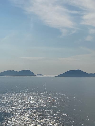 The image shows a scenic view of islands in the ocean, with a bright, hazy sky above. The water reflects the sunlight, creating a shimmering effect on the surface. The islands are silhouetted against the horizon, with a soft, diffused light illuminating the scene. The sky is a pale blue, with wispy white clouds scattered across it. This scene appears to be a typical seascape found in coastal areas, and could potentially be observed from a boat or a coastal viewpoint. Without additional context, it is impossible to definitively determine the specific location in Nam Nha Trang, Vietnam, or if the image was taken there at all.