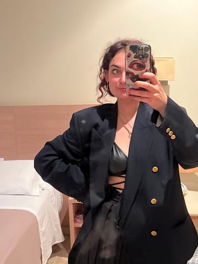 A person is taking a mirror selfie in a room. The person is wearing a black blazer with gold buttons and a black top. The phone case is patterned. The room appears to be a bedroom, with a bed and headboard visible. The location is Puçol, Spain.