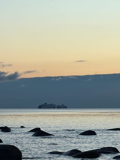 Two large passenger ferries are docked on the calm sea, silhouetted against a hazy sky of pale yellow and blue. In the foreground, dark, wet rocks emerge from the gentle waves of the Baltic Sea near the coast of Haabneeme alevik, Estonia. The image captures a serene moment, likely at dawn or dusk, with minimal lighting and a muted color palette. There are no people visible in the frame, and the ferries appear stationary. The scene is tranquil and devoid of any activity.