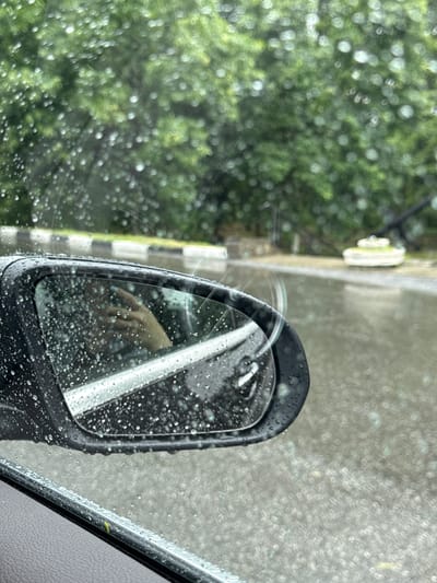 The image depicts a view from inside a vehicle during rainfall, looking towards the side mirror and the exterior. The side mirror is covered in raindrops, reflecting a person's hand holding a phone. Outside, the road is wet and appears to be flooded in some areas. Trees with green foliage are visible in the background, blurred by the rain. A sidewalk with black and white markings is also present along the road. In the distance, a round structure with white detailing sits on the wet ground. The overall environment suggests a rainy day in Oktyabrskoye, Russia, possibly on a street or in a park area given the presence of trees and a decorative structure.