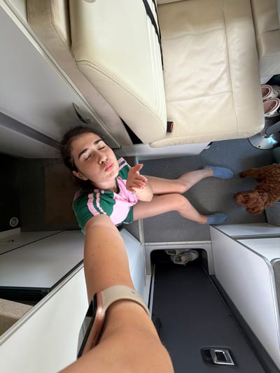 The image shows a young woman and a dog inside a vehicle, possibly a camper van or RV. The woman is taking a selfie, puckering her lips as if to blow a kiss. She is wearing a pink and green t-shirt and blue slipper socks. The dog, a small, curly-haired breed, is lying on the floor next to her. The interior of the vehicle appears to be upholstered in light-colored leather and has a practical, utilitarian design. There are storage compartments visible, one of which contains a pair of shoes. The flooring is a dark gray speckled material. The image is shot from a high angle, looking down on the woman and dog. The scene is well-lit, suggesting it is daytime. The overall impression is one of a casual, indoor setting within a vehicle. There is no visible indication of the city of Nea Moudania, Greece, in this image.
