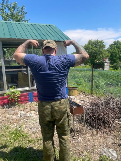 An outdoor, eye-level, medium shot captures a Caucasian man, viewed from behind, flexing his biceps under a bright blue sky with scattered white clouds. He wears a camouflage baseball cap, a dark blue t-shirt with a small black logo on the back, and camouflage cargo pants. His arms are covered in colorful tattoos.
The man stands on a gravelly area in front of a red-sided structure with a green, corrugated metal roof. To his left is a greenhouse-like structure with glass panels. Behind him, a wire fence encloses a green field of crops, with a rustic wooden building visible in the distance. A rusty barbecue grill with burning embers sits to the right of the man, accompanied by a large pile of dry branches and twigs. The overall setting suggests a rural or suburban environment, possibly a backyard or garden area. The scene is well-lit by natural sunlight, indicating a daytime shot.