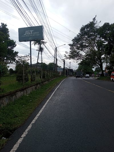 The image captures a street scene in Kamji Na-Yekha, Bhutan, under an overcast sky. A dark asphalt road, marked by a faded white line on its left edge, extends towards the horizon, curving slightly to the right. To the immediate left, a low concrete wall supports a metal fence, enclosing an overgrown, grassy field containing sparse vegetation and trees. Prominently positioned above the fence line is a large, rectangular dark green billboard displaying "J&T CARGO" in white uppercase letters, mounted on a tall metal pole. Numerous utility poles line the road, supporting a dense matrix of overhead electrical and communication wires that crisscross the sky. Several trees with full green foliage, including tall palm-like structures and broadleaf trees, are visible on both sides of the road. Further down the road, on the right side, commercial buildings are present, one featuring a partially legible blue sign. Vehicular traffic includes a dark-colored truck, a motorcycle, a white passenger car, and a red and white van, all traveling in the same direction away from the viewer. No specific individuals are discernible.