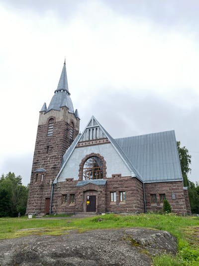 The image shows a stone church with a tall, octagonal steeple on the left. The church is constructed from rough-hewn, reddish-brown stones. It has a gabled roof made of corrugated metal. A large, arched window with wooden mullions is visible on the front facade, above the entrance. The entrance consists of two dark wooden doors. The church is situated on a grassy knoll, with a large boulder in the foreground. Trees and other greenery are visible on the right and left sides of the building, and in the background. The sky is overcast and a pale grey. A single person in dark clothing is visible walking on the grass to the left of the church. A sign with Cyrillic text is mounted on the exterior wall of the church to the right of the entrance. This is the church of St. John the Baptist, located in Melnikovo, Russia.