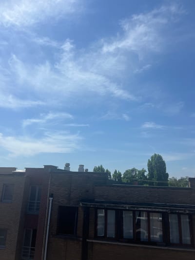 The image shows a view of a brick building against a bright blue sky with wispy white clouds. The building appears to be several stories tall and has numerous windows. Some of the windows are dark, suggesting they are not lit from within, while others appear to reflect the sky. The roofline of the building is visible, with several ventilation units or pipes protruding from it. Trees line the top of the building, suggesting an urban or suburban setting. The sky is clear and bright, indicating it is daytime, likely midday given the lighting. There are no people visible in the image. The overall scene conveys a sense of an ordinary day, with the focus on the architecture and the sky. The image is oriented vertically, making it appear as if the viewer is looking up at the sky and the building. The lighting suggests a sunny day.