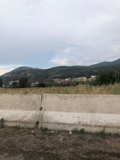 This media file depicts a mountainous landscape with some buildings. The scene is oriented vertically, suggesting the image was taken with a phone held sideways.
The primary subjects are the sloping, tree-covered mountains that dominate the background. Nestled within the mountains, and visible along the hillside, are several multi-story buildings, appearing to be residential or hotel complexes. The foreground is dominated by a cracked concrete structure, possibly a roadside barrier or a stepped pathway. To the right of the concrete structure is a patch of dry, rough ground with scattered stones and sparse vegetation.
The time of day appears to be late afternoon or early evening, given the soft, diffused light and the hazy sky. The sky itself is mostly overcast with some lighter patches indicating thin clouds. The overall mood is serene and somewhat distant, with no people or animals visible.
While specific location cues like street signs are absent, the context provided places this scene in Khujakent, Uzbekistan, which aligns with the mountainous terrain and architectural style of the buildings. There is no visible text in the image.