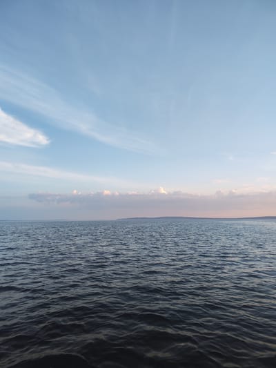 The image depicts a wide expanse of dark, choppy water under a pale blue sky. Wispy white clouds are scattered across the upper portion of the sky, with a band of soft pink and white clouds closer to the horizon. A low, dark landmass is visible in the far distance, forming the horizon line. The water exhibits numerous small waves, suggesting a gentle breeze. The overall scene conveys a sense of open space and tranquility. The location is identified as the city of Galevo, Russia. There are no visible people, specific objects, or discernible actions taking place in the image.