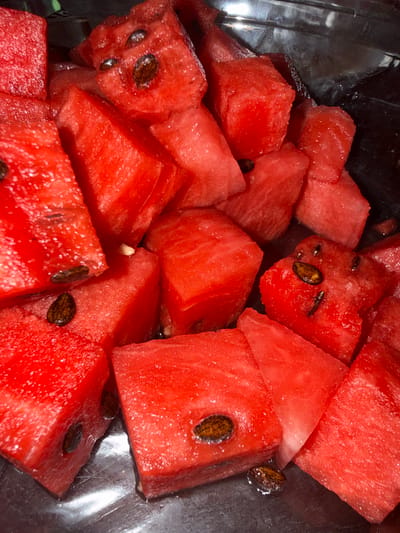 The image is a close-up, overhead shot of cubed watermelon in a dark, metallic container. The watermelon pieces are bright red and appear juicy, with some seeds visible. The container's surface is reflective, showing some glare and dark areas, suggesting it might be stainless steel or a similar material. The lighting is somewhat dim, creating shadows and highlighting the texture of the fruit. There is no visible text. The scene depicts a simple presentation of prepared fruit, likely ready for consumption. The focus is entirely on the watermelon and its textures, colors, and seeds. There are no people or other objects visible, and no context clues to suggest a specific time of day, weather, or precise location beyond being in Amstelveen, Netherlands.