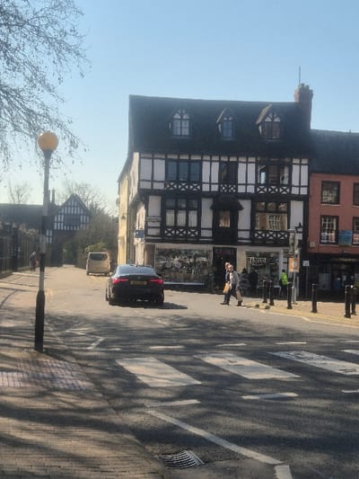 The image captures a daytime street scene on Broad Street in Hereford, United Kingdom, specifically looking south towards Bridge Street and Cathedral Close.
The central feature is a multi-story, black and white timber-framed building (26 Broad Street / 1 Bridge Street) with a dark, gabled roof and three dormer windows. Its ground floor houses commercial premises, including "Sarah J Hollinshead - Florist." To its immediate right, a terracotta-hued building is partially visible. In the background on the left, a smaller, traditional black and white timber-framed structure, characteristic of Cathedral Close architecture, is discernible.
On the asphalt road in the mid-ground, a dark-colored BMW sedan (F30 3-series, UK registration beginning 'YG...') is positioned, facing away from the camera. Further down the road, a light-colored van is also moving away. A clearly marked zebra crossing with white stripes occupies the foreground.
Two individuals are observed crossing the street on the zebra crossing, moving from right to left. One person wears a light jacket and dark trousers, carrying a light-colored bag. A second individual is partially obscured behind them. On the right-hand pavement, a person in a high-visibility yellow/green jacket stands near a series of dark bollards. A single individual walks on the left-hand pavement towards the background structures.
The environment is illuminated by bright sunlight under a clear blue sky. Leafless tree branches are visible in the upper left, casting distinct shadows across the left pavement and road. A black streetlamp with a yellow Belisha beacon is on the left pavement, and a drain cover is present on the right pavement edge.