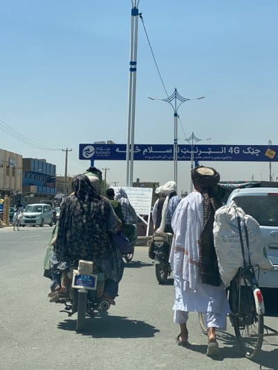 The image depicts a street scene in Kandahar, Afghanistan. Several people are visible, some on motorcycles and one walking with a bicycle loaded with a large sack labeled "Al-Khair." The individuals are wearing traditional clothing. Signage is visible in the background written in Arabic script and Persian, advertising "4G Internet." There is a sign showing the logo of "Salaam." Modern streetlights are visible. The scene suggests a daytime urban environment.