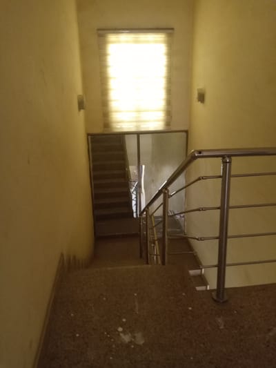 This media file depicts an interior view of a stairwell. The walls are painted a light yellow, and a bright, horizontally slatted window is visible at the top of the stairwell, letting in ample light, suggesting it is daytime. On either side of the window, simple wall sconces are mounted. The stairs themselves are made of dark, speckled material, likely granite or terrazzo, and appear to be leading downwards. A modern stainless steel handrail with multiple horizontal bars lines the right side of the stairwell, curving with the stairs. To the left of the stairs, a wall continues downwards. A glass partition separates the main stairwell area from a section of stairs that appears to be further down and to the left, also with a dark surface. The lighting creates a somewhat stark and functional atmosphere. There are a few white splotches on the stairs in the foreground, possibly paint splatters. No people or specific activities are visible. The location context provided is Hotoro North, Nigeria.