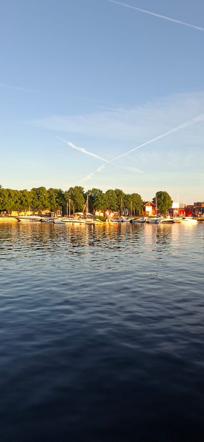 This is a vertical, eye-level, outdoor, daylight, full shot photograph taken from the water. The foreground is dominated by dark blue water with ripples reflecting the sky. In the midground, a harbor scene unfolds with numerous sailboats and motorboats moored along a wooden dock. Behind the boats, a line of lush green trees forms the backdrop, with several red-roofed buildings interspersed among them. The sky above is a clear, pale blue, with two distinct white contrails forming an 'X' shape. The lighting suggests it's late afternoon or early evening, with the sun casting a warm glow on the scene. The location appears to be a marina or harbor in Pärnu linn, Estonia, given the visual characteristics of the architecture and natural environment. There are no people visible in the image.