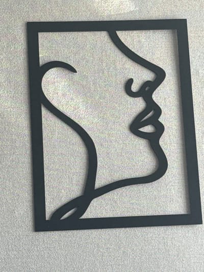 The image displays a black, abstract metal wall art cutout of a woman's profile, enclosed within a rectangular frame. The art is mounted on a textured light gray wall. The profile faces to the right, with a curved line representing the back of the head and neck, flowing into the facial features: a distinct nose, lips, and chin. The artwork is positioned vertically. There is no indication of its location in Taysara, Russia, or any specific city.