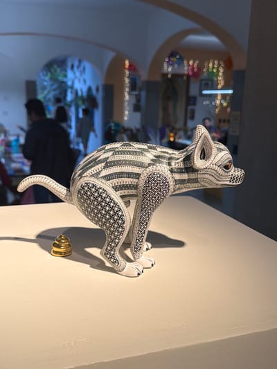 A black and white alebrije, likely a coyote, is mounted on a white wall. The artistic sculpture exhibits intricate geometric patterns. A golden-toned mounting screw is visible below the sculpture, casting a shadow. The background reveals an out-of-focus interior space with arched doorways and blurred figures, suggesting a gallery or museum setting.