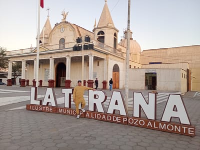 The image shows a man posing in front of a large sign that reads "LATRANA ILUSTRE MUNICIPALIDAD DE POZO ALMONTE", indicating he is in the city of Pozo Almonte, Chile. The man is wearing a mustard-colored tracksuit, a baseball cap, and sneakers. He is leaning against the "T" of "LATRANA" with one leg slightly raised. In the background is a large, white church with multiple towers and a statue on top. There are other people walking in the background, and a small building is to the right of the church. The ground is paved with hexagonal tiles. The sky is a clear, light blue.