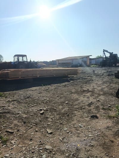This media file depicts an outdoor scene at a lumber yard, likely in Strelka, Russia. The setting is a dusty, outdoor area with piles of lumber, a tractor with a front loader, and a crane truck. In the foreground, a large stack of finished lumber is visible, resting on wooden supports. Behind the lumber, a blue tractor is parked, facing left. Further in the background, a long, low building with a wooden roof stands, with a crane truck parked to its right. The sky is a clear, bright blue, with the sun shining intensely and creating lens flare. The time of day appears to be midday or early afternoon, given the bright sunlight and strong shadows. The ground is rough and unpaved, covered with dirt, small stones, and some scattered wooden debris. The overall impression is one of a working industrial or agricultural site on a sunny day. There are no people visible in the image.