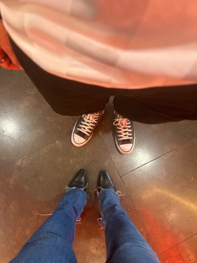 An overhead perspective depicts the lower bodies of two individuals standing on a dark, possibly concrete or tiled, floor exhibiting visible seam lines. In the foreground, one person wears blue denim jeans with visibly frayed lower hems and black, pointed-toe ankle boots. Directly behind this individual, a second person is positioned, wearing black trousers and black high-top canvas sneakers with white laces and toe caps. A portion of a white garment and a ring on a finger are discernible in the upper left of the frame, belonging to the second individual. The floor surface displays a reddish-orange ambient illumination, particularly concentrated on the right side of the image. The specific location within Leganés, Spain, is not identifiable from the visual information presented.