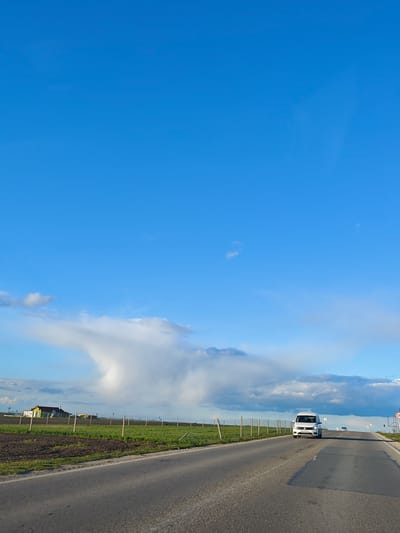 A wide shot captures a rural or semi-rural road scene under a bright, late afternoon sky. The dominant feature is a vast, clear blue sky occupying the upper two-thirds of the image, with scattered faint white clouds. A particularly striking, large white cloud formation, resembling a mushroom or anvil cloud, dominates the mid-ground on the left, contrasting with the expansive blue.

An asphalt road stretches from the foreground into the distance, showing signs of wear with several dark patches. A white van, possibly a Volkswagen Caddy, is prominently featured in the mid-ground, traveling towards the viewer. Another smaller, light-colored vehicle is visible further down the road in the distance.

To the left of the road lies a cultivated field, exhibiting patches of dark tilled earth and vibrant green vegetation. A simple fence line, composed of wooden posts and wire, runs along the left side of the road, separating it from the field. In the far distance on the left, behind the field, a yellow house with a reddish roof is discernible, accompanied by other smaller structures. On the right side of the road in the distance, a blue and white circular sign indicates a speed limit of "50".

The lighting suggests late afternoon or early evening, casting gentle light on the scene and highlighting the cloud formations. The overall impression is one of open space and travel through the Bulgarian countryside near Kichevo.