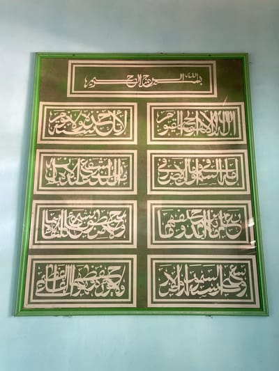 A framed Islamic calligraphy artwork hangs on a light blue wall. The artwork features a green background with a green frame. The calligraphy is in white, stylized Arabic script. The artwork is divided into several sections. The top section has a larger inscription, and below it are two columns of smaller inscriptions, with three inscriptions in each column, totaling six. The entire piece is mounted on the wall. The image appears to have been taken from a straight-on perspective, with the artwork filling most of the frame. The location provided is Sekar Mawar, Indonesia.
