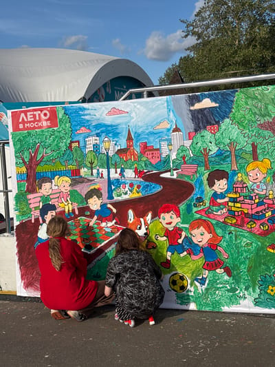 The image shows a large, colorful mural being painted outdoors. The mural depicts a park scene with children playing various games. The mural is on a white banner, and at the top left corner, there is text that reads "ЛЕТО В МОСКВЕ", which translates to "SUMMER IN MOSCOW". Two women are kneeling and painting the mural, one wearing a red shirt and the other a black and white patterned shirt. The background of the mural features trees, buildings, a cityscape, and a blue sky with clouds. The scene appears to be a public event or activity.