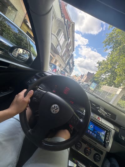 The image is a first-person perspective from inside a Volkswagen car, driving down a city street. The driver's hand is visible on the steering wheel, which has a Volkswagen logo in the center. The dashboard features a digital infotainment screen displaying various icons, including navigation, music, and apps. The car's interior is dark, contrasting with the bright, sunny exterior.
Outside the car, a street lined with buildings and trees is visible. The architecture of the buildings suggests a European city. The sky is blue with scattered white clouds. In the distance, other cars are seen on the road. A side mirror reflects another car.
The text on a poster in the distance, "ockyvziel," is a clue that this image was taken in Waregem, Belgium. The overall impression is that of a scenic drive through a European city on a pleasant day.