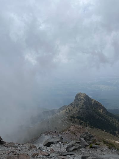 The image depicts a mountainous landscape partially obscured by heavy fog or low-hanging clouds, possibly in the city of Guadaloupe, Mexico, or nearby its environs. The foreground shows a steep, rocky cliff face with large boulders and visible vegetation. Further up the slope, small groups of people can be seen hiking. The terrain is rugged and slopes downwards. In the background, through the fog, there are indications of a distant city or populated area, marked by flat terrain and structures. The sky is overcast and uniformly gray.