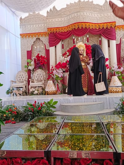 This image depicts a wedding ceremony taking place in Meuliek, Indonesia. The bride, adorned in traditional attire and a golden headdress, stands at the center of a decorated stage. She is flanked by two women in black hijabs, one of whom appears to be adjusting the bride's veil or dress. The stage is elaborately decorated with red and gold drapery, ornate carved panels, and abundant floral arrangements featuring red roses and other colorful blooms. Two ornate white chairs are positioned on the stage. In the foreground, a section of a reflective floor, possibly made of glass, is visible, mirroring the lights and decorations of the event. The overall atmosphere suggests a formal and festive occasion.