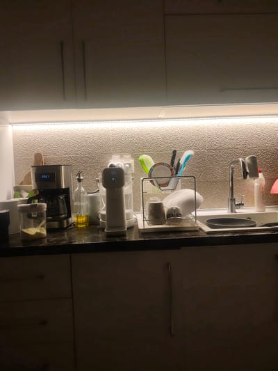 A well-lit kitchen counter features a coffee maker displaying the time "23:21", a clear glass bottle with yellow liquid, a white water dispenser with a digital display showing "85", and a dish rack filled with plates, cups, and utensils. A kitchen sink with a faucet is visible to the right, and below the counter, cabinets with handles are present. The environment appears to be indoors, likely a residential kitchen in Neapoli Municipal Unit, Greece, given the user's prompt.